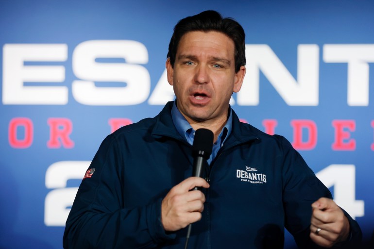 Governor Ron DeSantis speaks into a microphone in front of a screen that shows his presidential campaign logo.