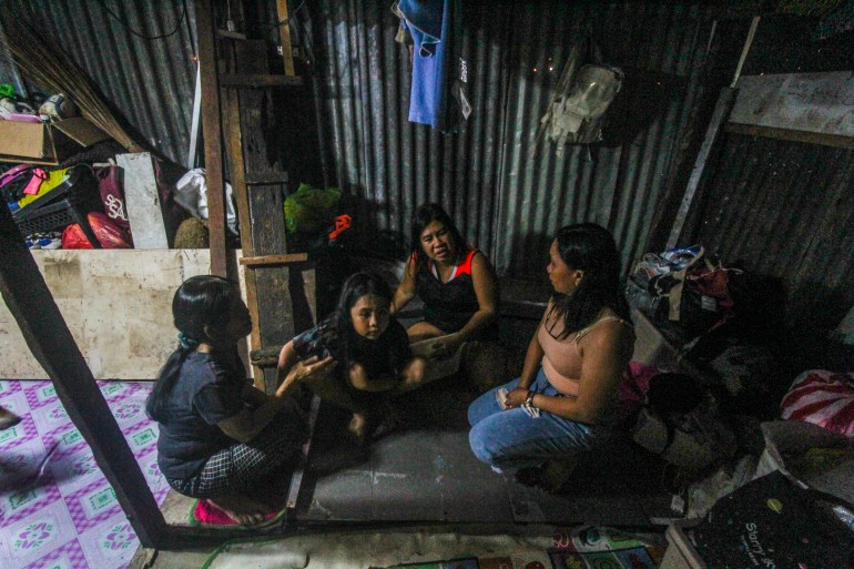 Merjelda Toralba sitting on the floor inside her home. She is with her family. It is quite dark. There are lots of belongings around them. The walls are made of sheets of corrugated iron.