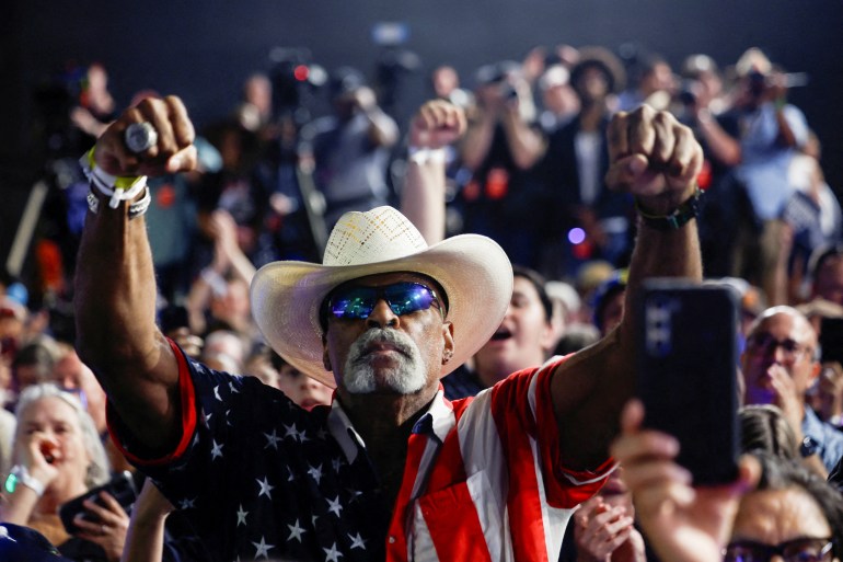 A supporter in a broad-brimmed hat, sunglasses, and a US flag shirt raises two fists for Kamala Harris, as a sign of support.