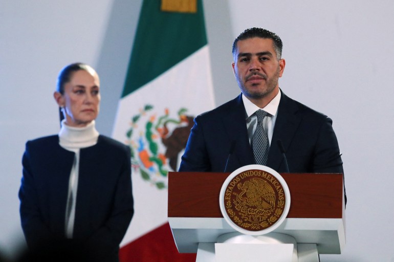 Omar Garcia Harfuch stands behind a podium speaking, while Claudia Sheinbaum looks on, standing next to a Mexican flag.