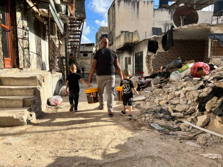 Man carries water on a tarmac road followed by his two children surrounded by rubble