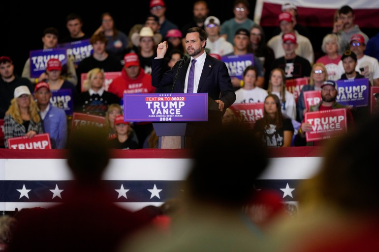 JD Vance stands behind a Trump-branded podium and speaks to a crowd.