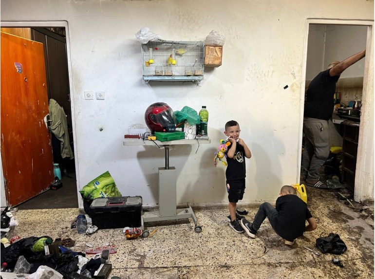 A bird cage is seen in the background as two children play in a partially destroyed room and their father make coffee in a small kitchen