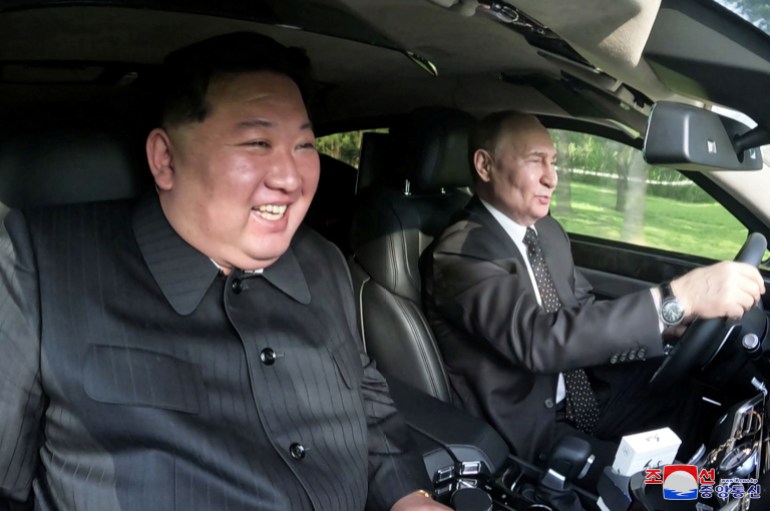 Kim Jong Un grinning as he sits in the passenger seat while Vladimir Putin drives the Aurus