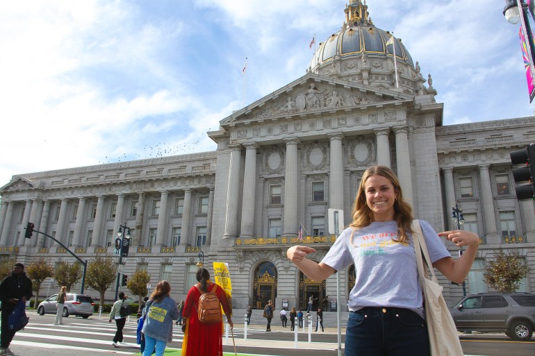 A woman points to her shirt in front of San Francisco City Hall. It reads: We are not going back