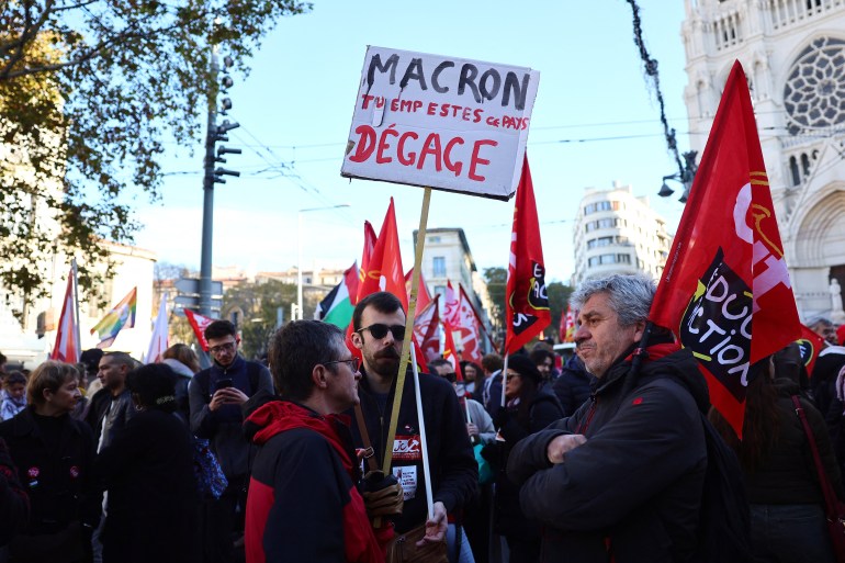 A protester holds a placard reading "(French President) Macron, you stink, get out" during a rally in Marseille on December 5, 2024, as part of a day of action and strike in the public sector. - Civil servants in France are mobilizing on December 5, 2024 for a day of action and strikes called by public service unions to open up a social front in the midst of a political crisis, the day after Michel Barnier's government fell on a motion of no-confidence.