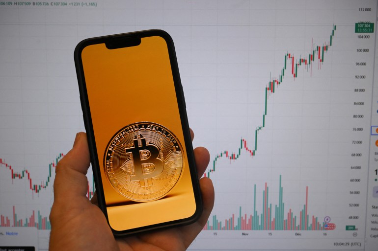 This illustration photograph shows a Bitcoin cryptocurrency coin picture displayed on a smartphone beside a screen showing a trading chart in Brussels on December 17, 2024. (Photo by Nicolas TUCAT / AFP)